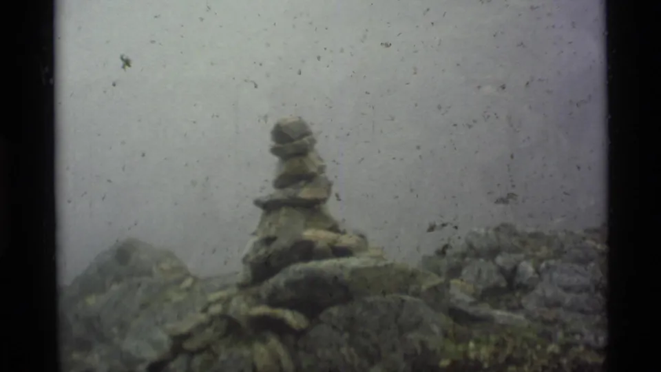 1977 Alaska 8mm frame showing a small stacked stone cairn amid wet rocks and mist, with visible dust and soft focus.