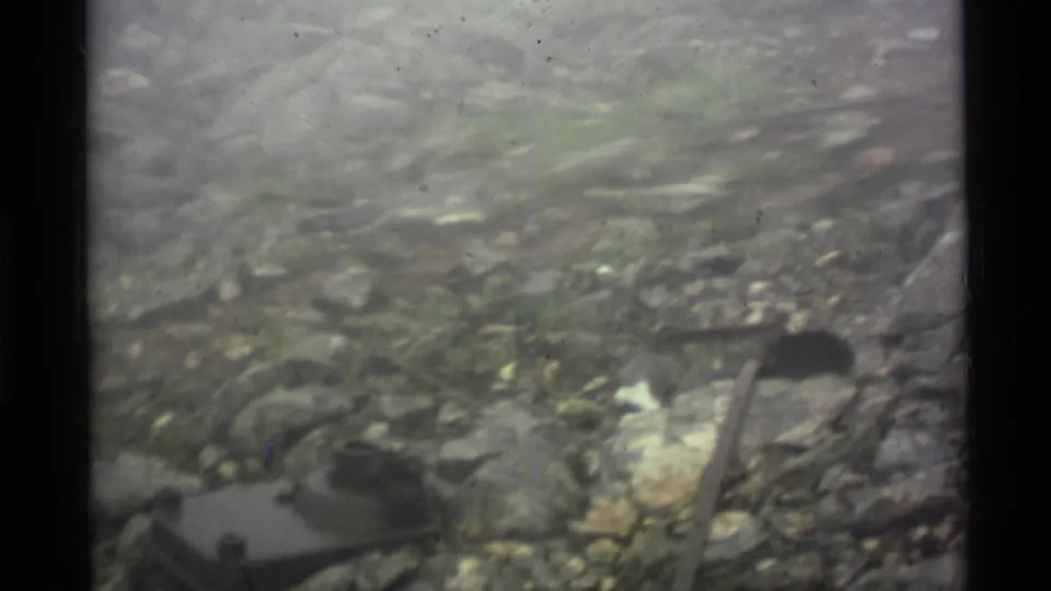 1977 Alaska 8mm frame showing a hammer on uneven rocky ground with roots, small debris, and soft gray outdoor light.
