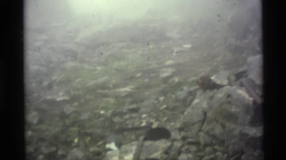 1977 Alaska 8mm frame with hazy rocky terrain, roots, scattered stones, and a partly visible hammer near the bottom of the frame.