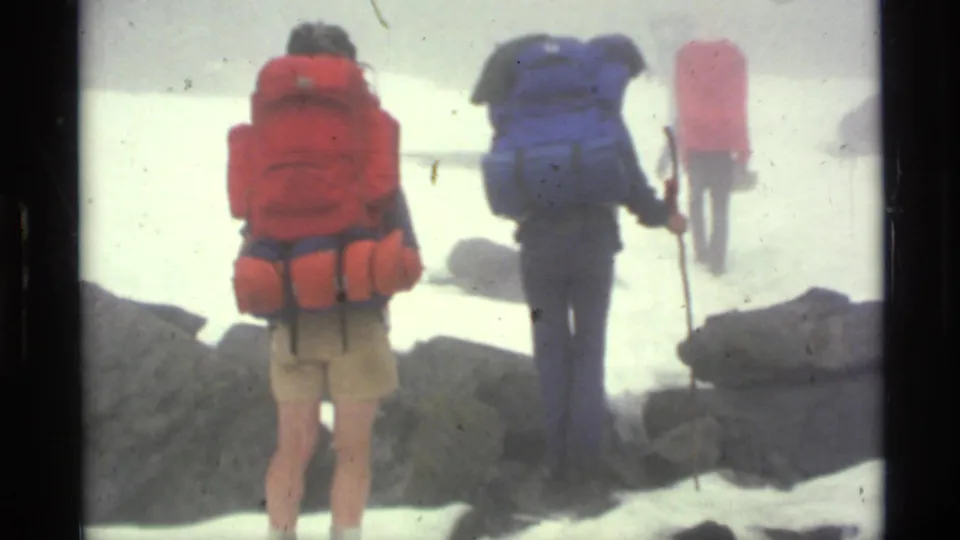 Three backpackers walk away through a snowy Alaskan pass, with red and blue packs prominent against foggy rocks.