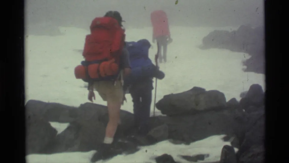 Rear view of backpackers navigating snow and boulders in an Alaska mountain pass in 1977 home-movie footage.