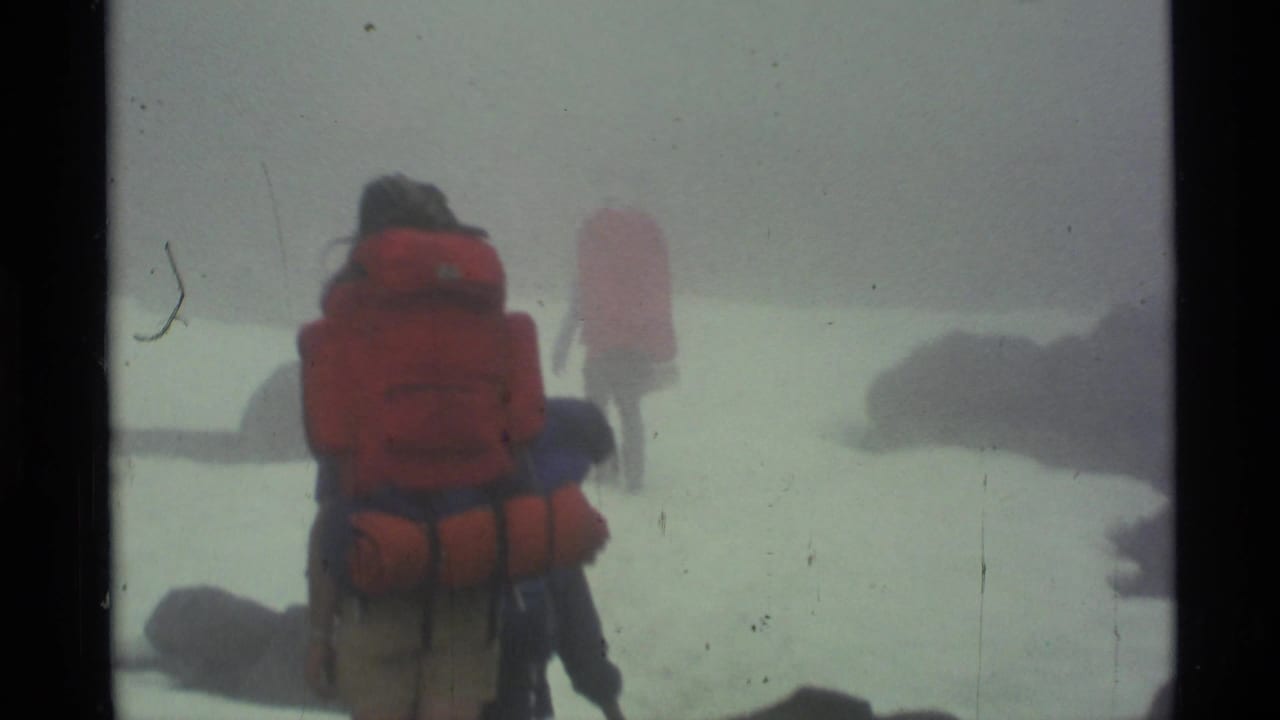 Hikers with red backpacks walk along a foggy rocky trail near water.