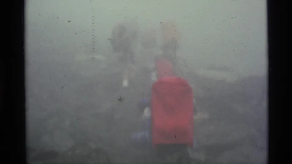 Fog-softened 1977 Alaska frame with a red-backpacked hiker and other figures partly obscured on rocky terrain.