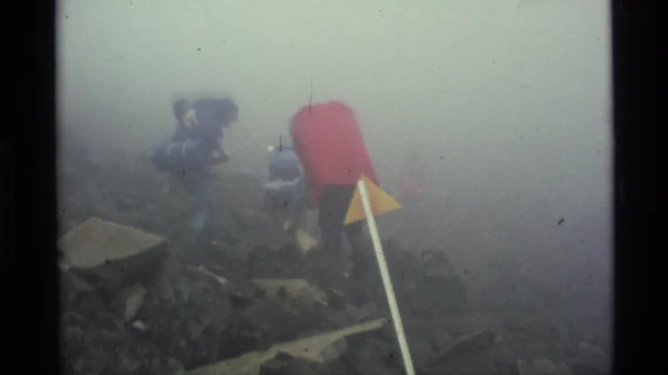 1977 Alaska 8mm frame of backpacked hikers moving away through fog across uneven rocky terrain.