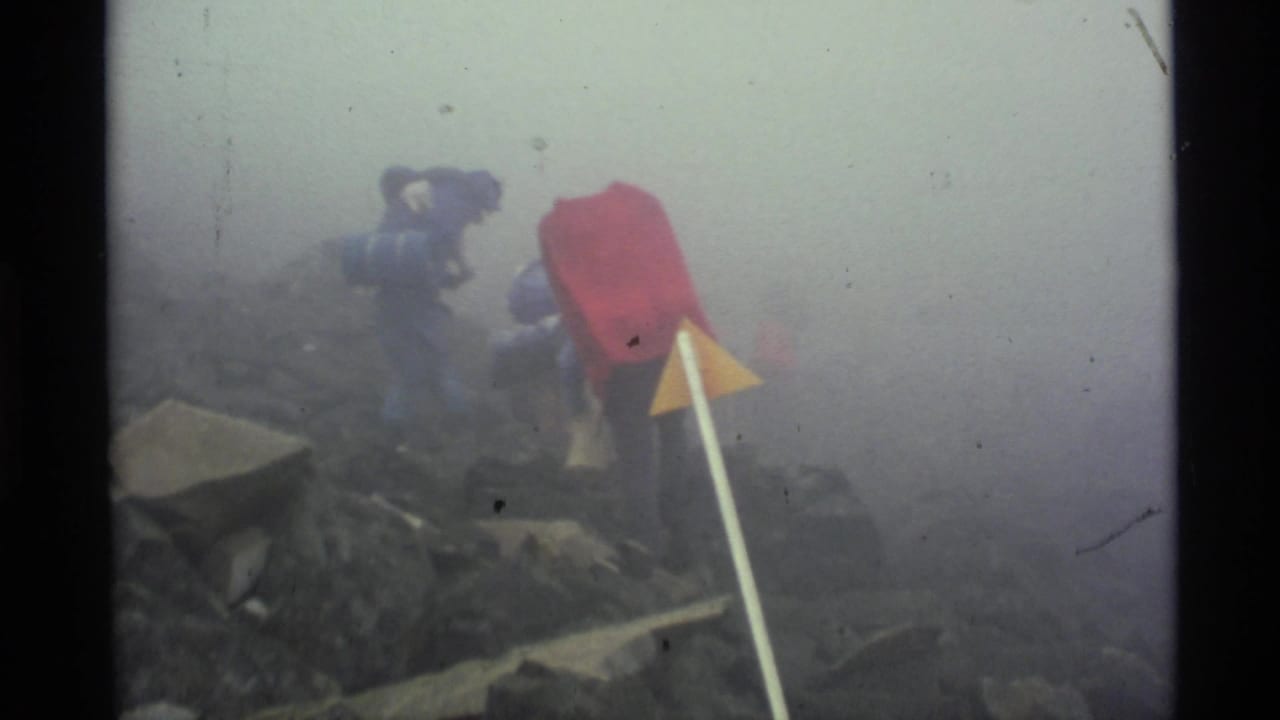 In 1977 Alaska home-movie footage, hikers with large packs move up a misty rocky slope near a yellow triangular trail marker.