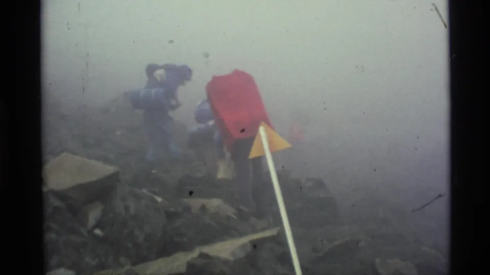 In 1977 Alaska home-movie footage, hikers with large packs move up a misty rocky slope near a yellow triangular trail marker.