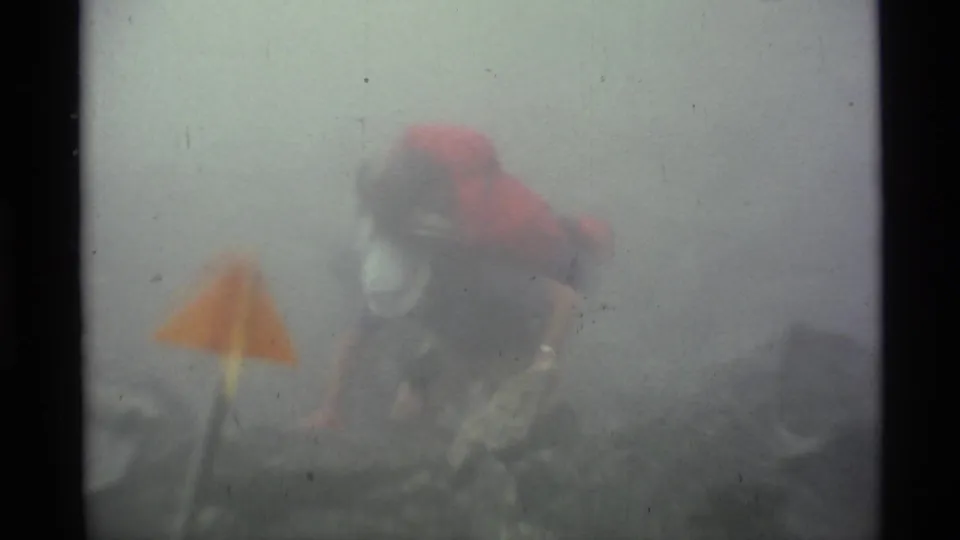 A backpacked hiker in a red jacket appears in thick Alaska fog near an orange trail flag on uneven mountain ground.