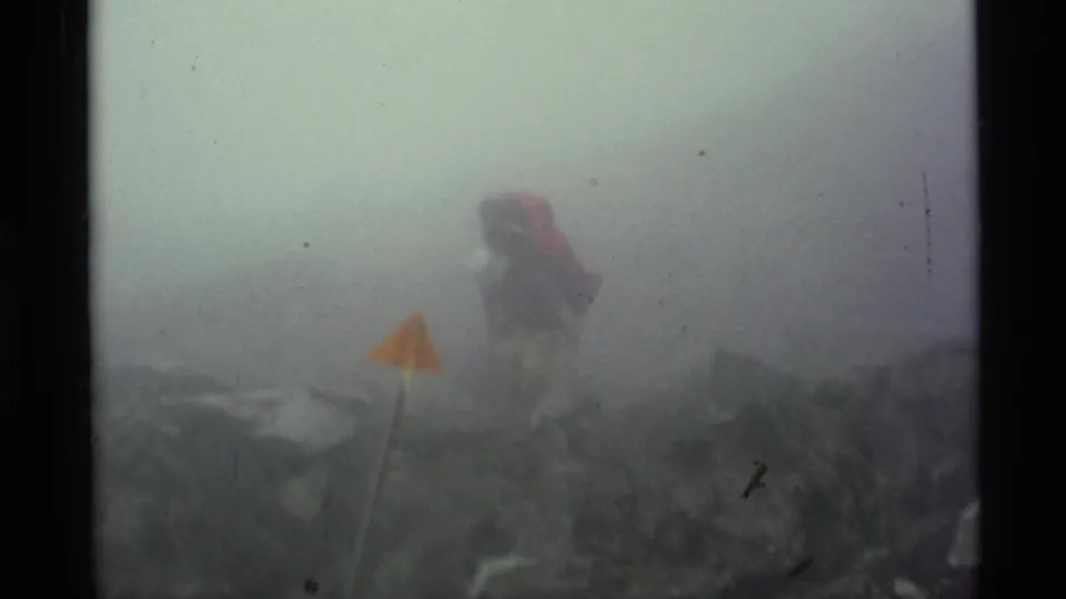 A distant hiker with a large pack is partly obscured by fog near a yellow-orange marker flag in 1977 Alaska footage.