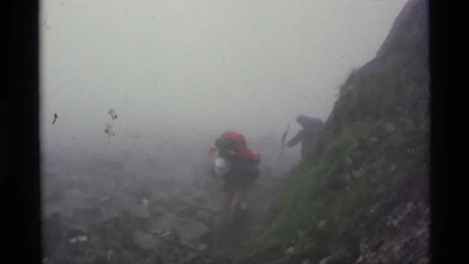 Distant hiker with a red backpack on a misty Alaska mountain trail in 1977 home-movie footage.