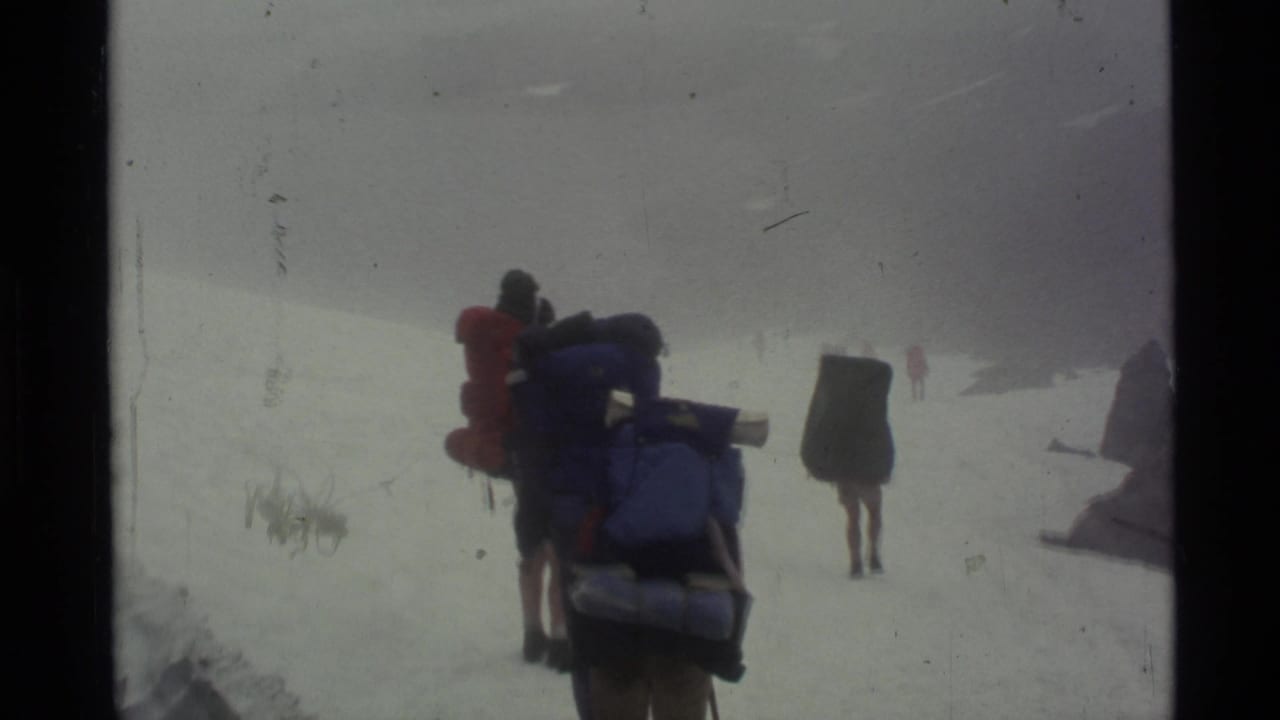 A loaded hiker follows others across snow in thick fog.