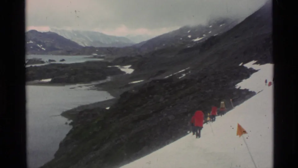 Wide 1977 Alaska 8mm view of hikers in red jackets on a snowy slope above a lake and rugged mountain terrain.