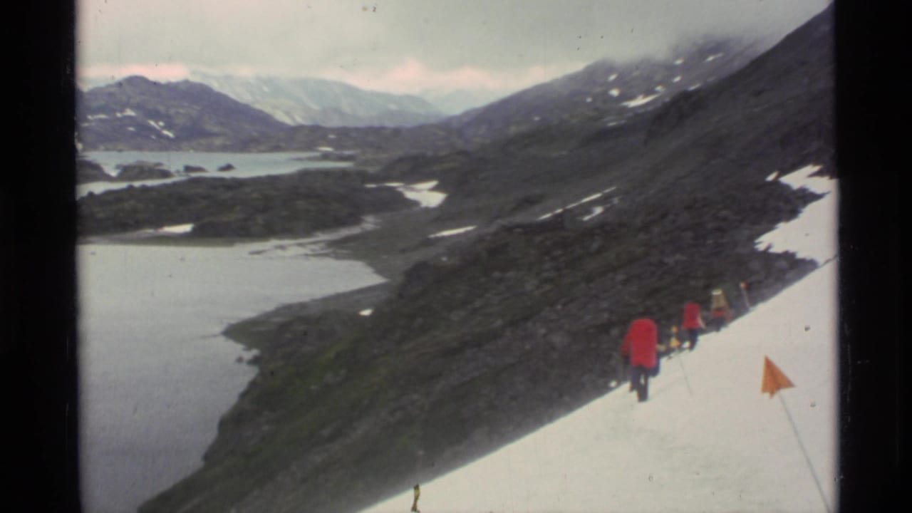 1977 Alaska 8mm frame showing red-coated hikers crossing a snow slope above glacial lakes and dark mountain ridges.