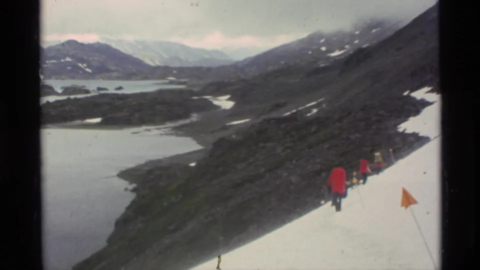 1977 Alaska 8mm frame showing red-coated hikers crossing a snow slope above glacial lakes and dark mountain ridges.