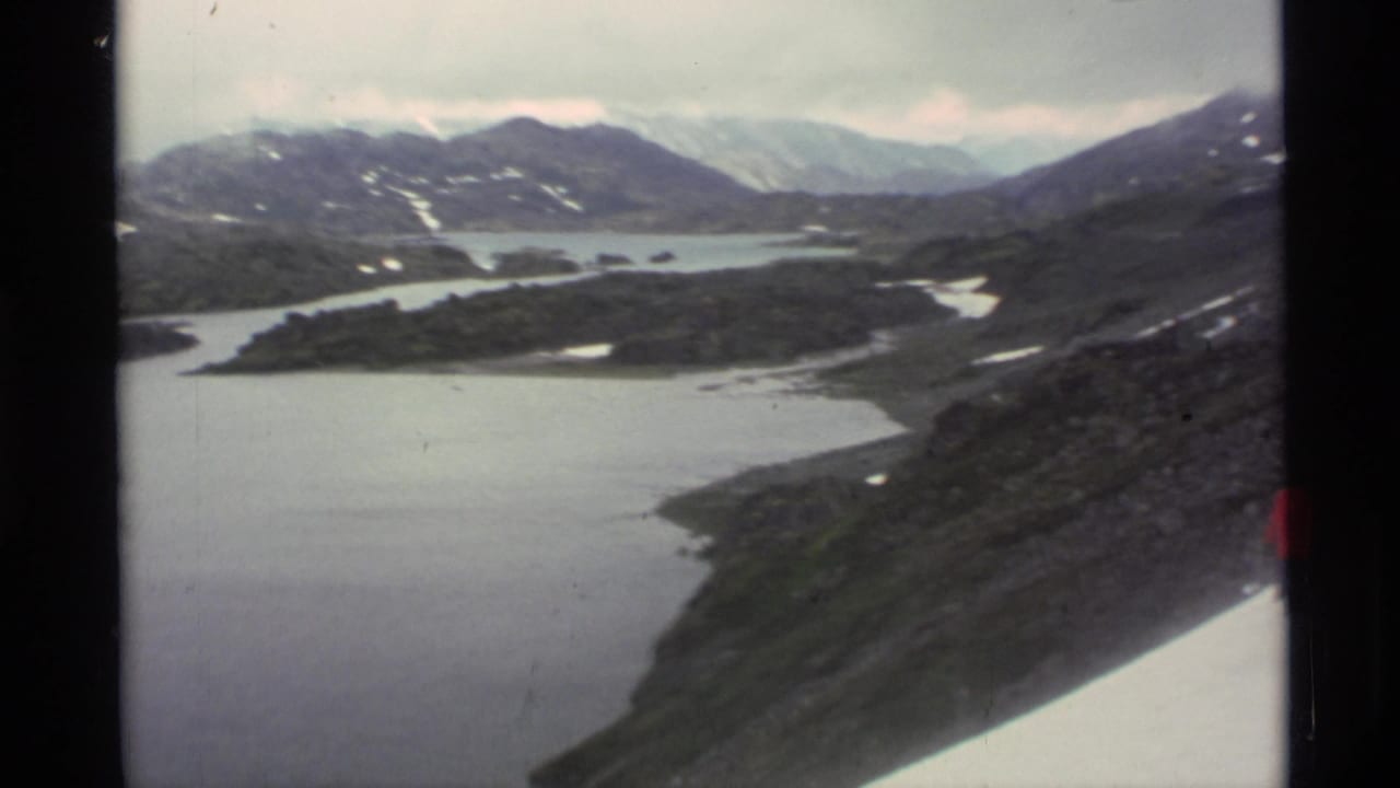 Wide mountain and lake view with a snowy slope at the right edge and a red figure partly visible.