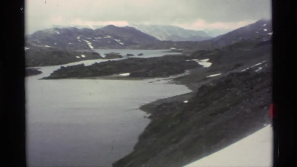 Wide 1977 Alaska 8mm view of lakes, rocky alpine terrain, distant mountains, and a partial red-coated hiker at frame edge.