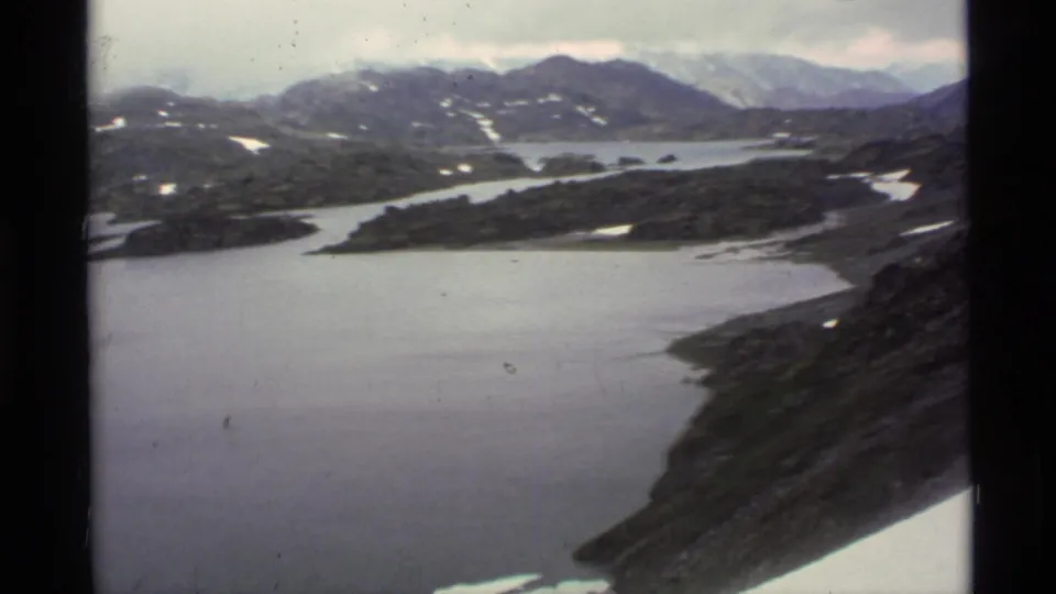 1977 Alaska 8mm frame looking over a glacial lake, dark ridges, snow patches, and distant mountain ranges.