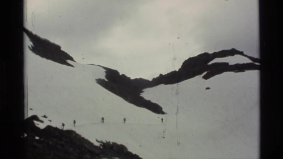 1977 Alaska 8mm frame with small hikers on a snowfield beneath dark exposed rock in an alpine mountain setting.