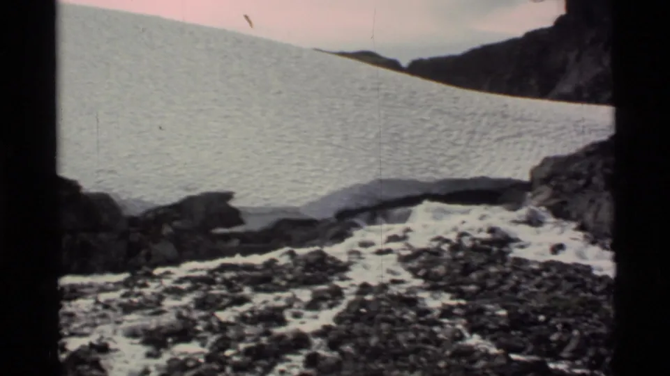 Wide 1977 Alaska mountain view with a broad snowfield, rocky foreground, and dark slopes at the edge of the frame.