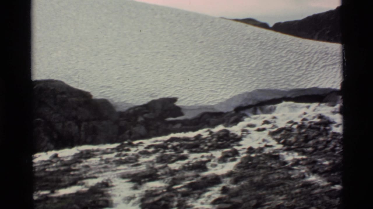 1977 Alaska 8mm frame showing a broad snowfield, dark rocks, and a rocky stream area below mountain slopes.