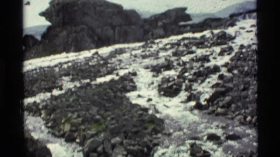 1977 Alaska home-movie frame with fast-moving water crossing a rocky slope beside lingering snow patches.