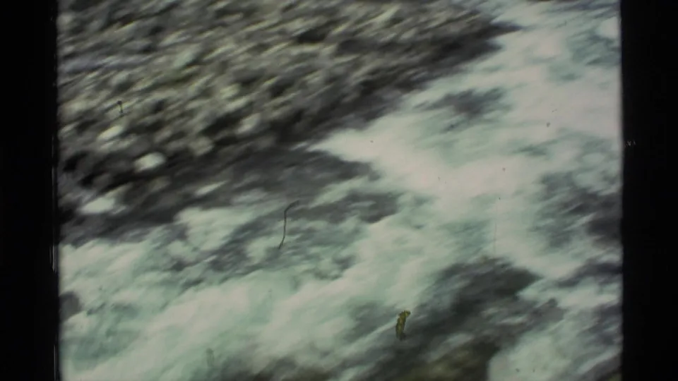 Soft, motion-blurred 1977 Alaska 8mm frame showing rushing water and dark rocks in a mountain stream.