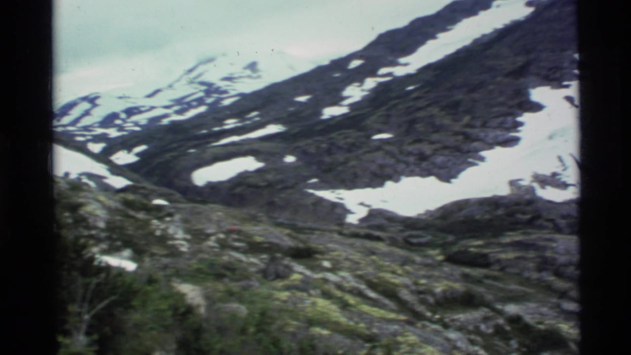 1977 Alaska 8mm frame showing a snow-dusted rocky mountain slope with green alpine vegetation in the foreground.