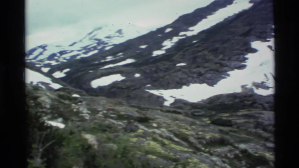 1977 Alaska 8mm frame showing a snow-dusted rocky mountain slope with green alpine vegetation in the foreground.