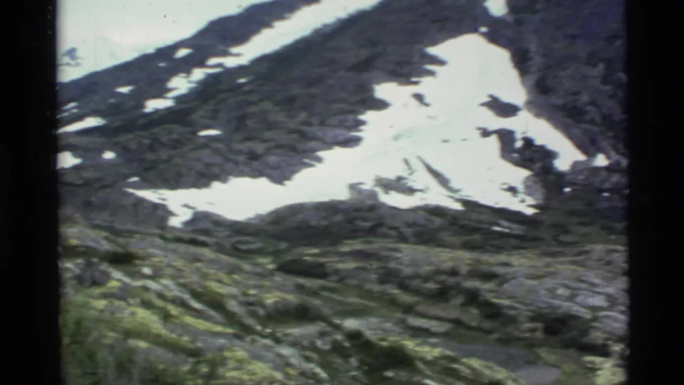 1977 Alaska home-movie frame of a rugged mountain hillside with white snow patches across dark rock and mossy ground below.