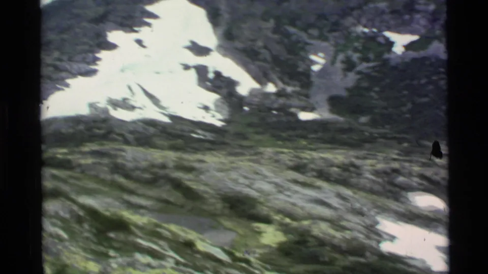 1977 Alaska 8mm frame looking across rocky, green alpine terrain toward a snow-marked mountain slope.