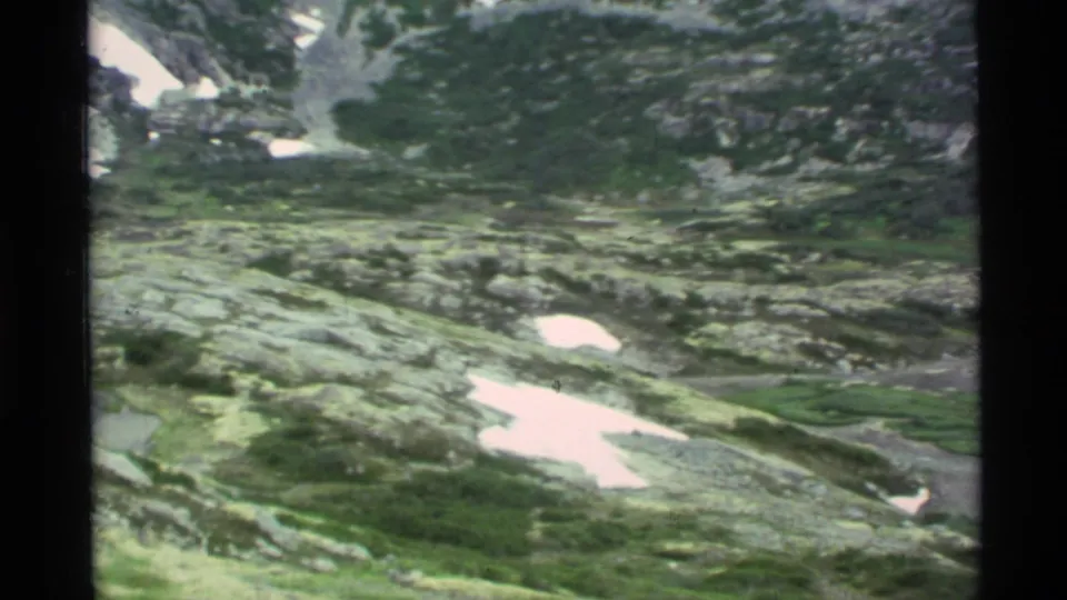1977 Alaska home-movie frame showing a broad rocky alpine basin with green vegetation and scattered white snow patches.