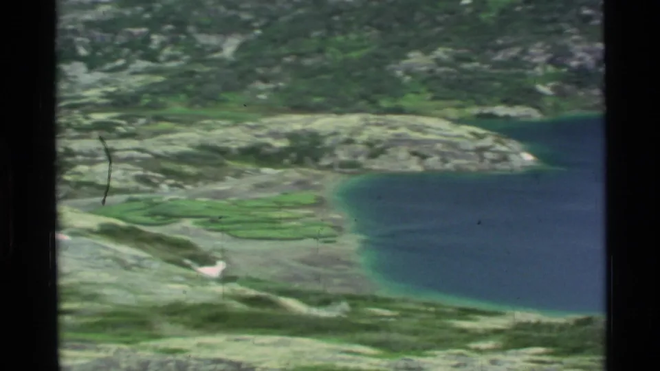 1977 Alaska home-movie frame showing a blue alpine lake edged by pale rocks, green ground cover, and turquoise shallow water.