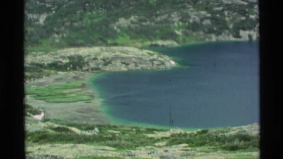 1977 Alaska 8mm frame with an elevated view of a blue lake, green shoreline flats, and surrounding rocky mountain terrain.