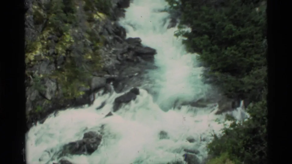 1977 Alaska home-movie frame showing a forceful waterfall cascading through dark rock and green forest.