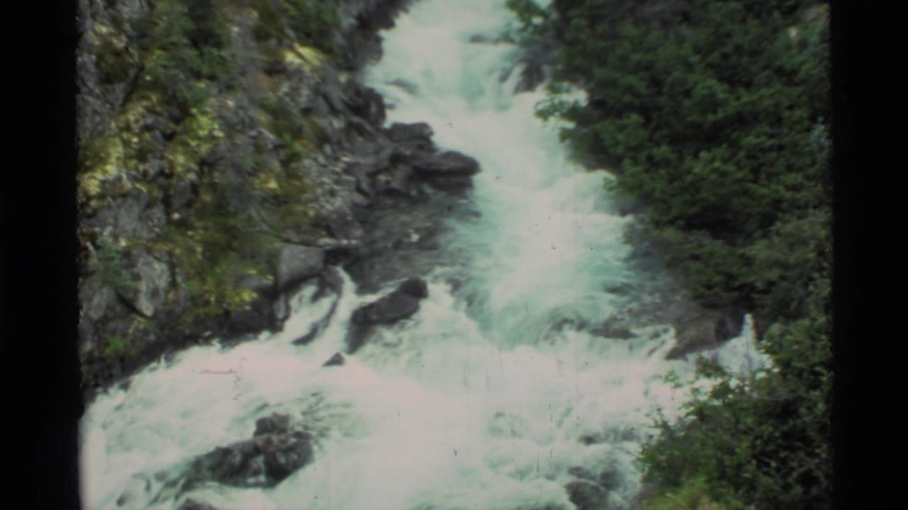 1977 8mm Alaska footage showing whitewater rapids churning through a rocky, tree-lined mountain channel.