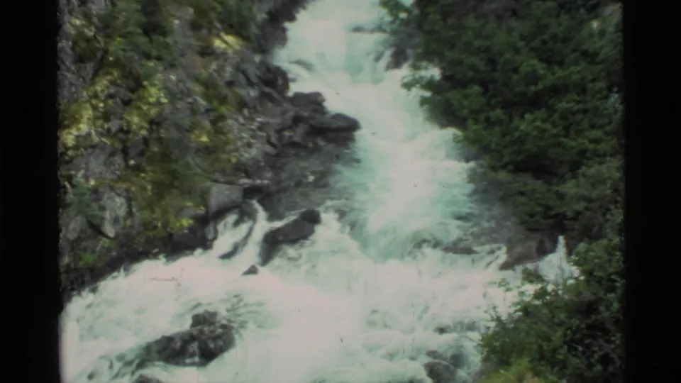 1977 8mm Alaska footage showing whitewater rapids churning through a rocky, tree-lined mountain channel.