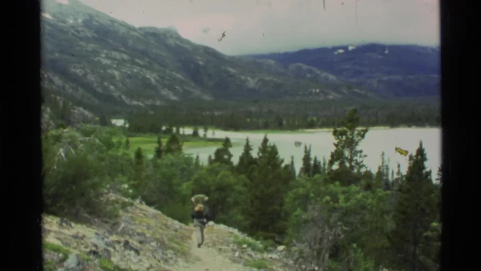 A distant backpacked hiker walks along an Alaska mountain trail overlooking a broad lake and evergreen valley in 1977 8mm footage.