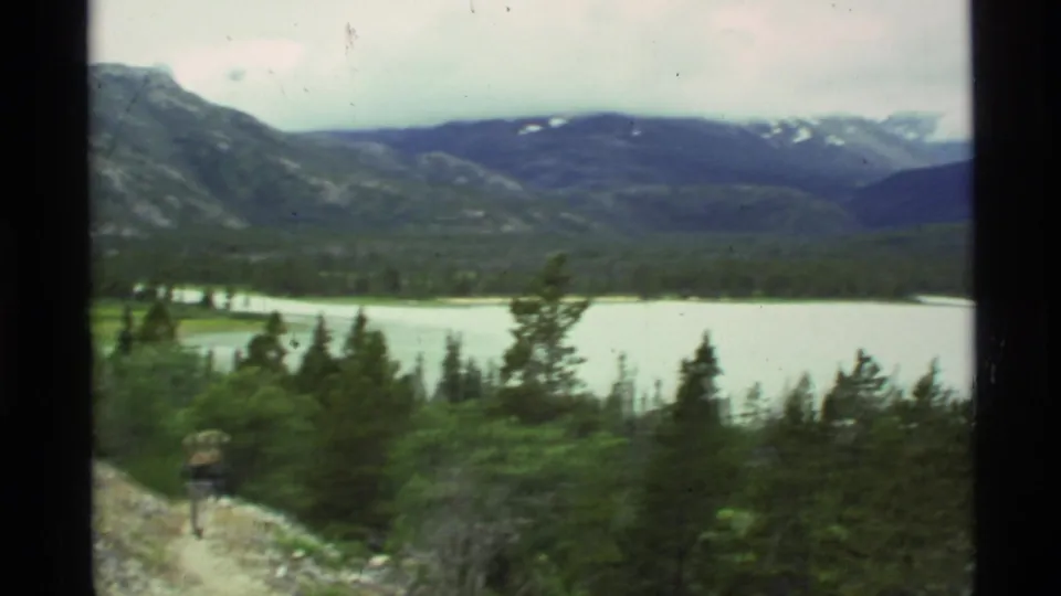 The backpacked figure appears at the edge of frame as the view opens to a lake, forest, and snow-dusted Alaska mountains.