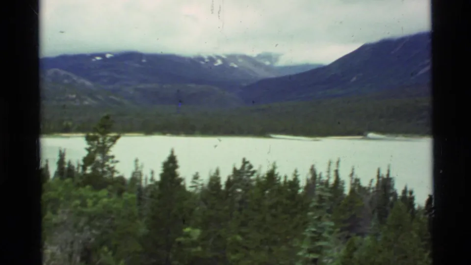 A wide 1977 8mm Alaska landscape frames a pale alpine lake, dense evergreens, and surrounding mountain ridges with snow patches.