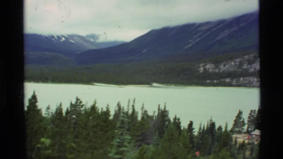 1977 Alaska 8mm landscape frame with evergreen treetops, pale lake water, rocky slope, and distant snow-dusted mountains.