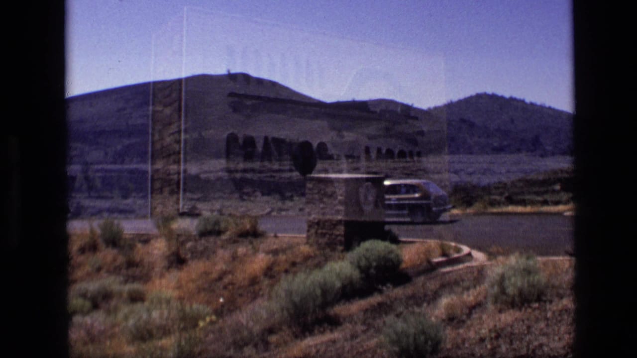 1972 Idaho 8mm frame showing a dark sedan near the Craters of the Moon sign, with stone walls, sagebrush, and hills behind it.