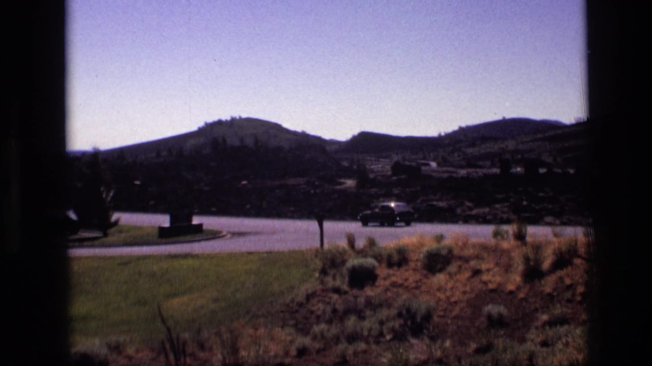1972 Idaho home-movie frame looking through a window toward a curved park road, open brush, distant hills, and a dark car.