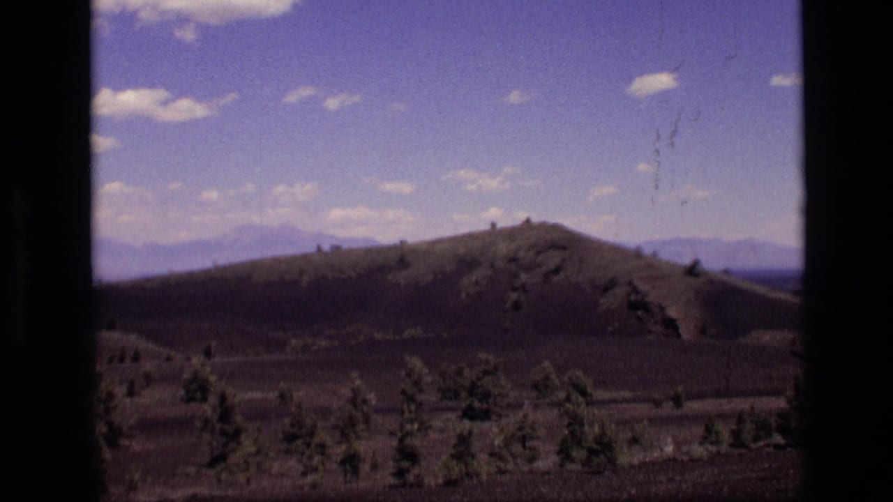 1972 Idaho 8mm frame showing a rounded hill, sparse evergreens, dark open ground, distant mountains, and a cloud-dotted blue sky.