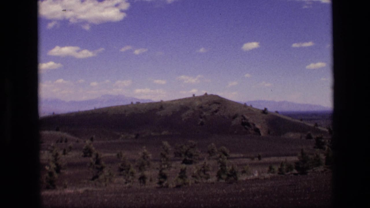 1972 Idaho home-movie frame with a low rounded hill, dark foreground plain, sparse trees, distant mountains, and blue sky.