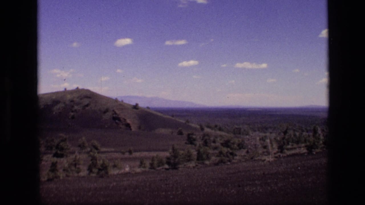 1972 Idaho home-movie frame showing rolling desert hills, sparse trees, distant ridges, and small clouds within dark film borders.