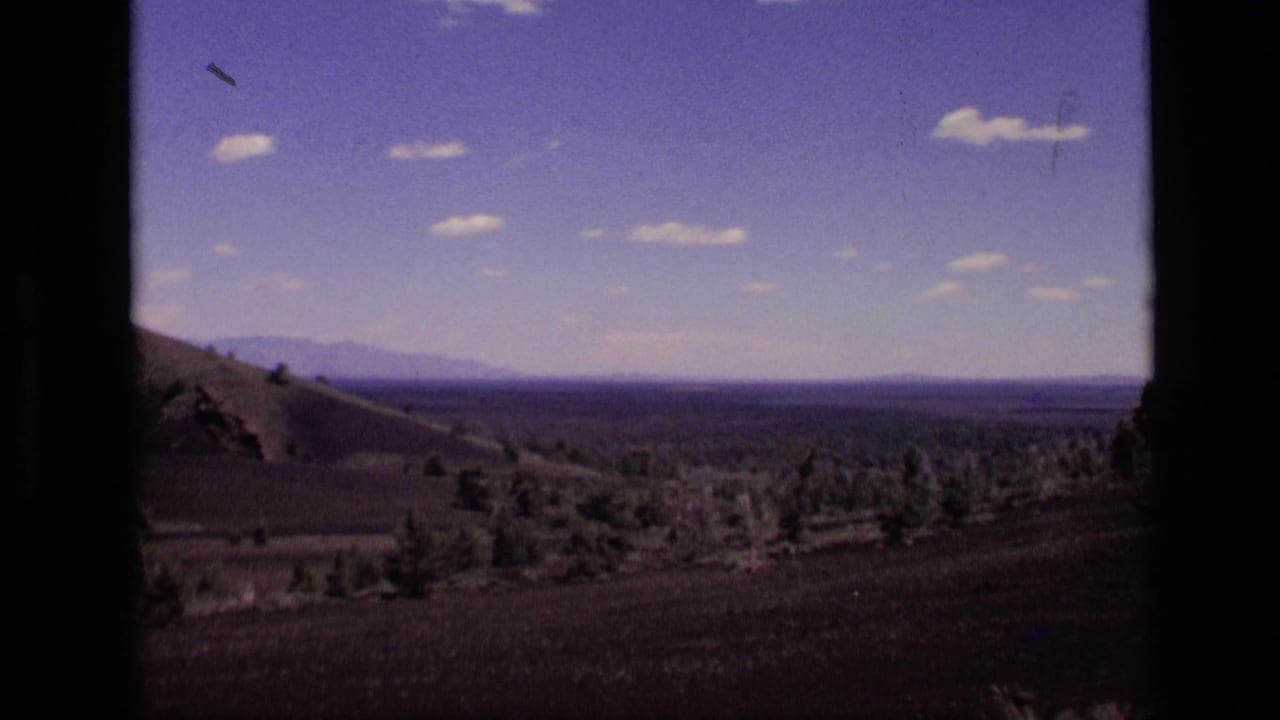1972 Idaho landscape view with a dry hillside at left, open valley, distant mountain ridge, and scattered clouds in a blue sky.