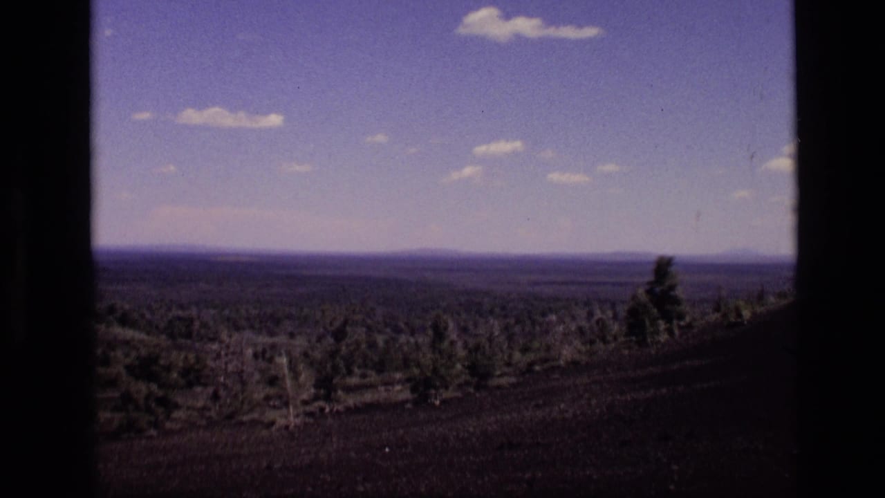 Wide valley landscape with distant mountains, sparse trees, blue sky, and film-frame borders.