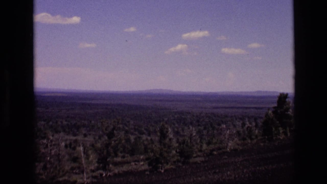 1972 Idaho home-movie view across a wooded foreground and open terrain toward a hazy mountain ridge beneath scattered clouds.