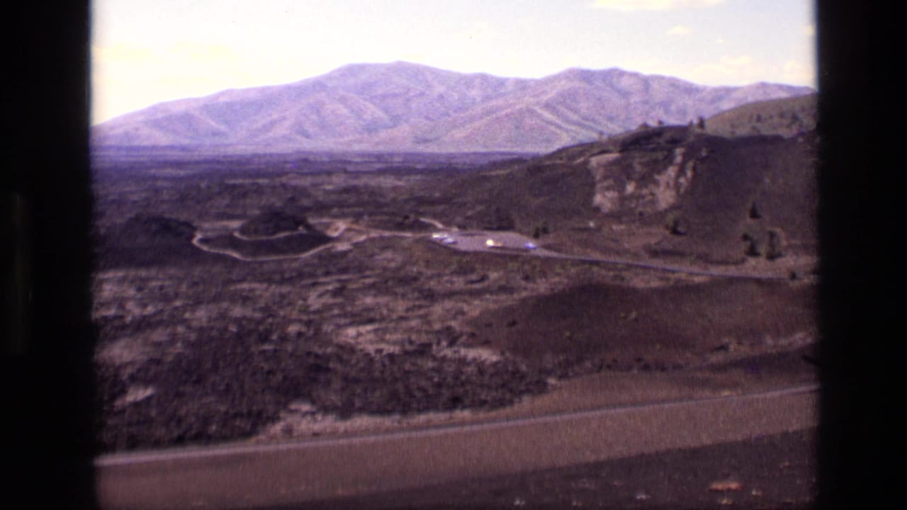 1972 Idaho 8mm frame showing dark rocky terrain, a distant mountain range, winding roads, and a small turnout under a pale sky.