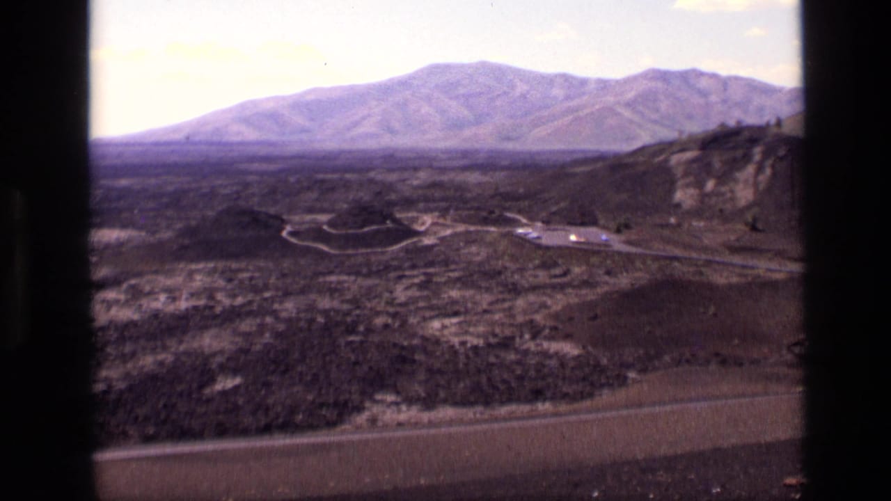 1972 Idaho home-movie frame with a broad mountain horizon, volcanic-looking ground, curving roads, and a small roadside turnout.