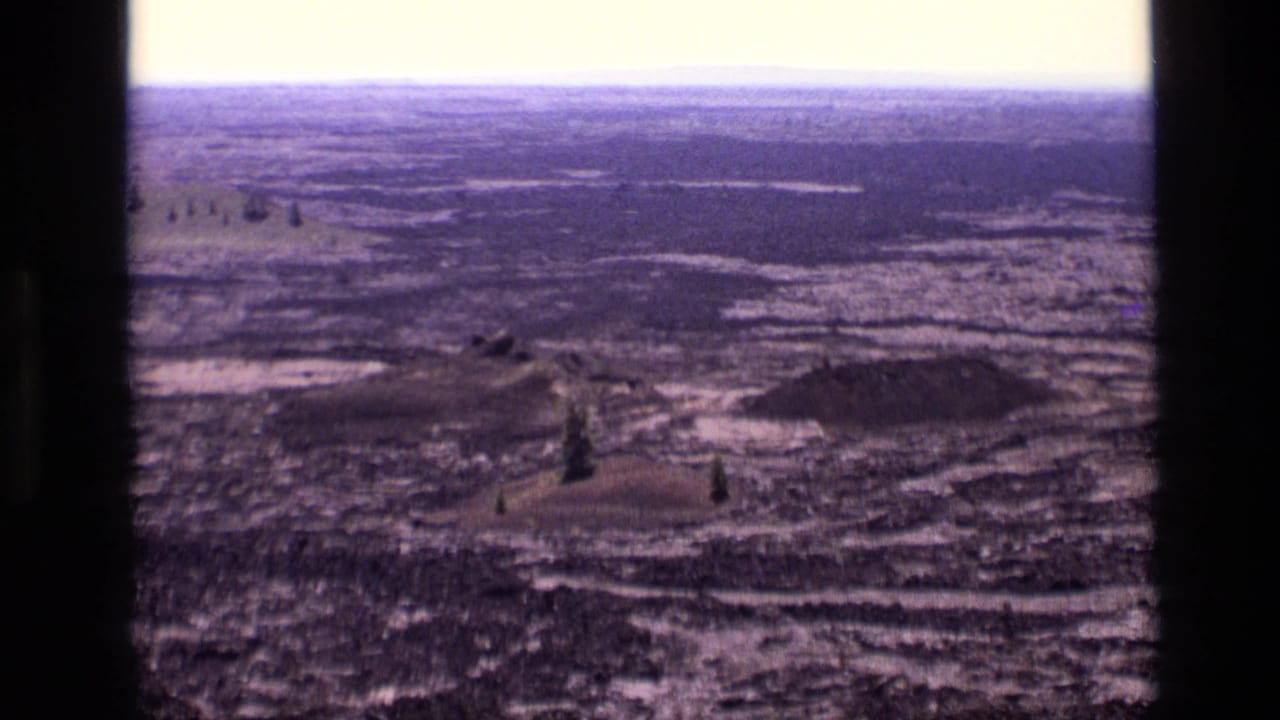1972 Idaho 8mm frame looking across dark rugged ground, scattered trees, and a distant horizon from a high overlook.